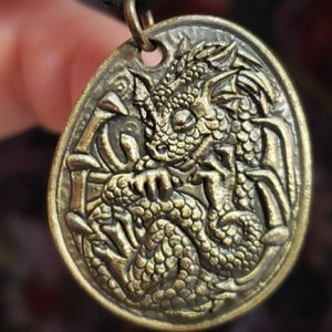 May include: A gold-toned metal pendant featuring a detailed dragon design. The dragon is curled up with its wings spread out. The pendant is attached to a black cord.