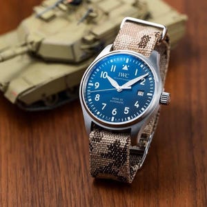 May include: A stainless steel IWC Mark XX automatic watch with a blue dial and a camouflage fabric strap. The watch has white hands and numerals, and a date window at the 3 o'clock position. The watch is set against a blurred background.