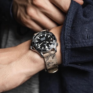 May include: A black-faced wristwatch with a silver bezel and a patterned tan and brown strap. The watch features white hour markers and hands, with a date display at the 3 o'clock position. The watch is secured on a wrist.
