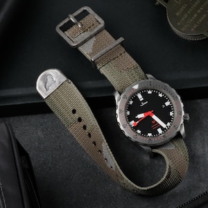 Puede incluir: Un reloj de pulsera con esfera negra, detalles en rojo y correa de camuflaje. La esfera muestra la marca "Sinn" y tiene agujas blancas. La correa es verde oliva y marrón con un patrón de camuflaje.