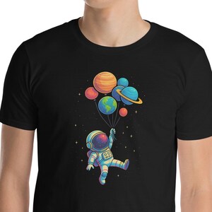Könnte beinhalten: Schwarzes T-Shirt mit einer farbenfrohen Illustration eines Astronauten, der mit Planetenballons schwebt. Der Astronaut trägt einen Raumanzug, und die Ballons stellen verschiedene Planeten dar. Das Design ist lebendig und skurril.