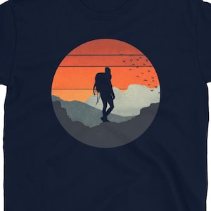 Puede incluir: Camiseta azul marino con un diseño gráfico circular de un excursionista silueteado contra un atardecer. El diseño incluye montañas, pájaros y tonos naranja, rojo y gris. La palabra "Navy" está impresa en blanco en la esquina superior izquierda.