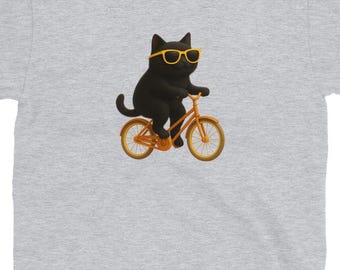 May include: Sport Grey t-shirt featuring a black cat wearing yellow sunglasses and riding an orange bicycle. The cat is the central focus, with the text "Sport Grey" at the top left. The shirt is a light gray color.