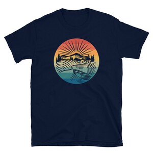 Pode incluir: Camiseta azul marinho com um gr&aacute;fico retro de um barco em um lago com montanhas e um p&ocirc;r do sol ao fundo.