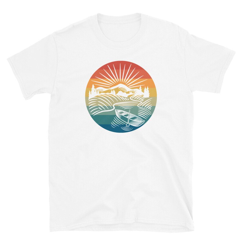 Pode incluir: Camiseta branca com um gr&aacute;fico de estilo vintage de um barco em um lago com montanhas ao fundo. O gr&aacute;fico est&aacute; em um c&iacute;rculo com um esquema de cores de p&ocirc;r do sol.