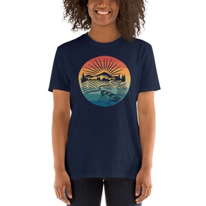 Pode incluir: Uma camiseta azul marinho com um gr&aacute;fico de estilo vintage de uma cena de lago com um barco, montanhas e um p&ocirc;r do sol. O gr&aacute;fico est&aacute; em um c&iacute;rculo com um degradado de cores arco-&iacute;ris.