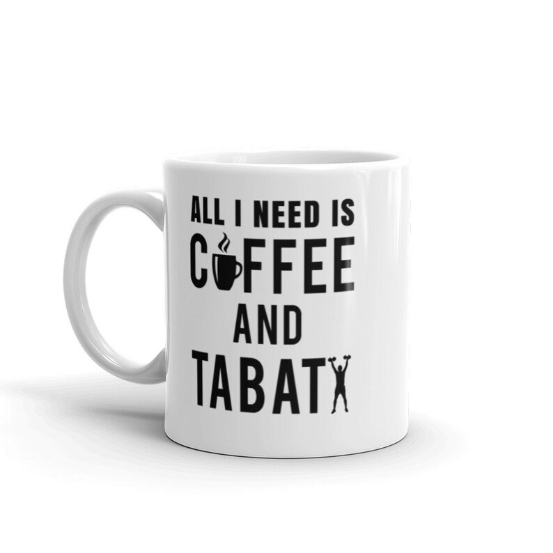 May include: White ceramic coffee mug with the text "ALL I NEED IS COFFEE AND TABATA" in black. The design includes a coffee cup icon and a person lifting weights. The mug has a curved handle.