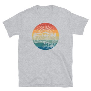 Pode incluir: Uma camiseta cinza com um gr&aacute;fico de estilo vintage de um lago com um barco e montanhas no fundo. O gr&aacute;fico est&aacute; em um c&iacute;rculo com um esquema de cores arco-&iacute;ris.