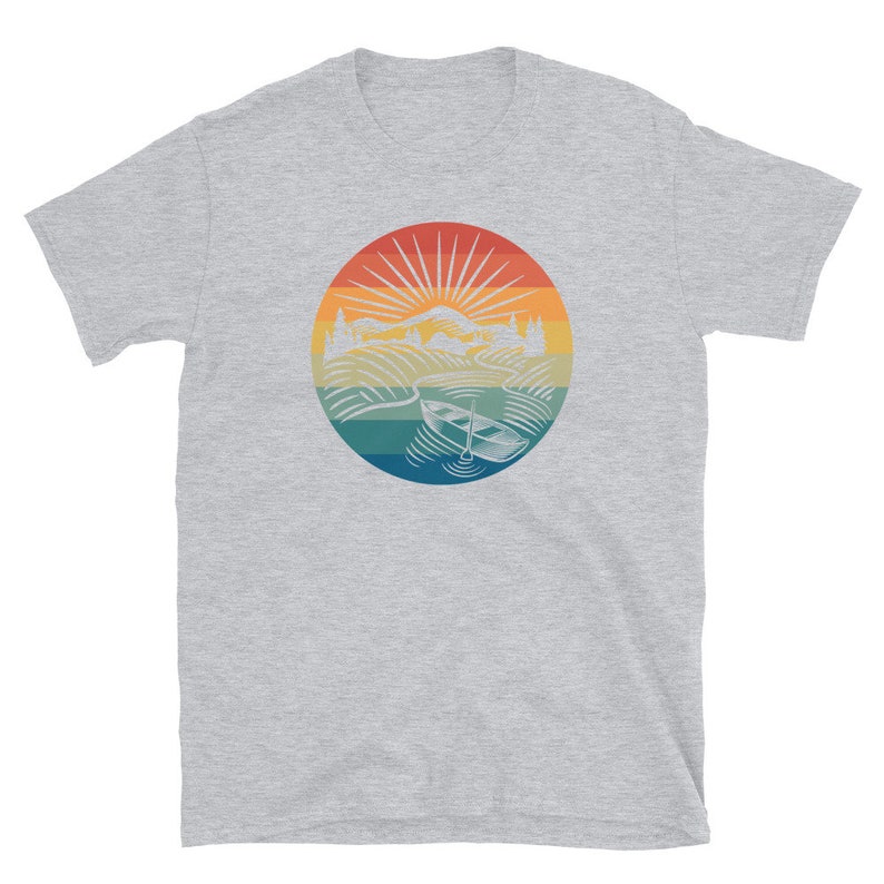 Pode incluir: Uma camiseta cinza com um gr&aacute;fico de estilo vintage de um lago com um barco e montanhas no fundo. O gr&aacute;fico est&aacute; em um c&iacute;rculo com um esquema de cores arco-&iacute;ris.