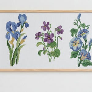 May include: A framed cross-stitch floral design featuring three distinct flower arrangements. The left side showcases blue irises, the center displays purple violets, and the right side presents blue morning glories. The artwork is set against a white background.