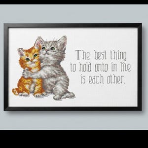 May include: Cross-stitch art in a black frame featuring two hugging kittens. One kitten is orange, the other is gray. Text reads: "The best thing to hold onto in life is each other."