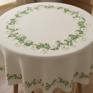May include: A round, off-white tablecloth with a daisy floral embroidery design. The embroidered daisies have white petals and yellow centers, with green leaves. The tablecloth is draped over a round table.