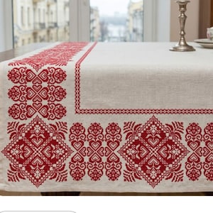 May include: A cream-colored table runner with intricate red embroidered patterns. The design features floral and geometric motifs along the edges and center, creating a decorative and traditional aesthetic. The runner is made of a natural fabric.
