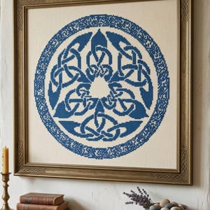 Op de afbeelding: Een ingelijst kruissteekkunstwerk met een blauw Keltisch knoopontwerp op een crèmekleurige achtergrond. Het kunstwerk wordt weergegeven in een goudkleurige houten lijst. Het ontwerp bevat een centrale knoop en een rand van kleinere knopen.