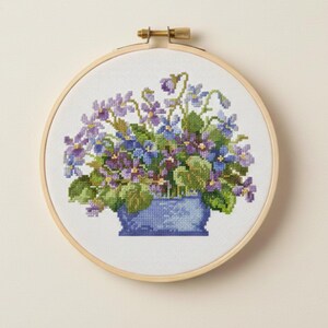 May include: A cross-stitch embroidery of a floral arrangement in a blue vase. The flowers are a mix of purple, blue, and green, set against a white background within a wooden embroidery hoop. The hoop is secured with a brass clasp.