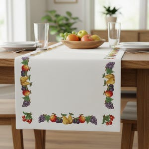 Op de afbeelding: Een witte tafelloper met een kleurrijke geborduurde rand met diverse vruchten. De loper ligt op een houten tafel, met een houten fruitschaal en glaswerk zichtbaar. Het ontwerp van de loper omvat druiven, peren en appels.
