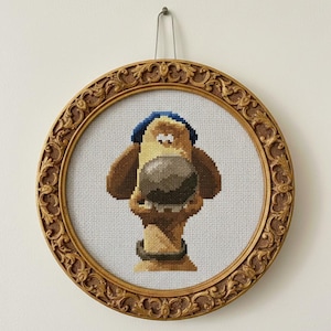 以下が含まれることがあります： 大きな鼻と青い帽子をかぶった漫画の犬が描かれた、額入りのクロスステッチアートワーク。犬は茶色、タン、グレーの色合いで白い背景にステッチされています。丸いフレームは、装飾的なディテールが施された木製です。