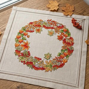 Könnte beinhalten: Ein quadratischer, beigefarbener Leinen-Tischläufer mit einem detaillierten gestickten Kranzdesign. Der Kranz zeigt Herbstblätter in Orange, Gelb und Grün, sowie rote Beeren und orangefarbene, laternenartige Blüten. Der Tischläufer hat einen genähten Rand.