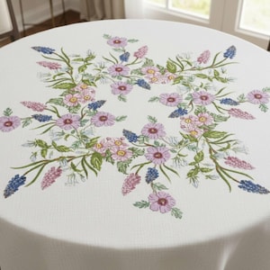 Può includere: Una tovaglia rotonda bianca con un motivo floreale. Il design presenta fiori rosa, viola e blu con foglie verdi disposte in un motivo circolare. La tovaglia è realizzata in un tessuto strutturato.