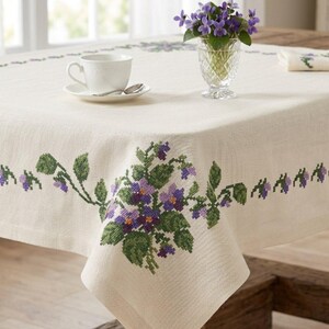 Peut inclure: Une nappe crème avec un motif floral brodé au point de croix représentant des violettes violettes et des feuilles vertes. Une tasse et une soucoupe blanches avec une cuillère sont sur la table, ainsi qu'un vase de violettes et une serviette assortie.