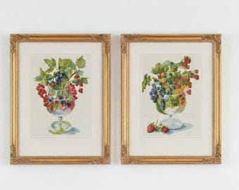 May include: Two framed cross-stitch pictures. Each depicts a stemmed glass filled with colourful berries and green leaves. The gold frames have ornate detailing. One glass has red and black berries, the other has red, purple, and yellow berries.