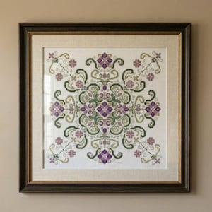 May include: Framed cross-stitch artwork with a symmetrical floral design in purple, green, and gold on a white background. The artwork is in a dark brown wooden frame with a gold inner border, creating a decorative wall hanging.