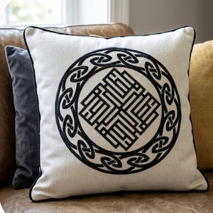 May include: Square decorative pillow with a cream-colored background and a black Celtic knot design. The pillow has a black rope trim around the edges. The pillow is on a brown leather couch.