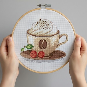 May include: Cross-stitch embroidery of a coffee cup with whipped cream, a coffee bean, and cherries. The embroidery is set in a wooden hoop. The design features shades of brown, beige, red, and green.