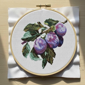 May include: Cross-stitch embroidery of a branch with plums. The plums are a mix of purple and pink hues, with green leaves. The embroidery is set in a wooden hoop, with white fabric surrounding it.