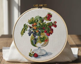 May include: Cross-stitch embroidery in a wooden hoop, depicting a glass filled with berries and grapes. The design includes red strawberries, purple grapes, and green leaves. Scissors and threads in various colours are on a wooden surface.