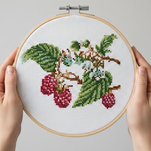 Puede incluir: Bordado de punto de cruz de una rama de frambuesa con hojas verdes y bayas rojas, en un bastidor de madera. La labor muestra puntadas detalladas, destacando la textura y las variaciones de color de la fruta y el follaje.