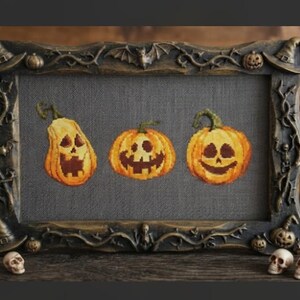 May include: A rectangular frame with a dark, weathered finish featuring sculpted bats, witch hats, and vines. Inside, a grey fabric panel displays three embroidered jack-o'-lanterns with distinct carved faces in shades of orange and yellow.