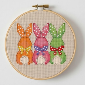May include: A cross-stitch embroidery hoop featuring three colorful bunnies. The bunnies are orange, pink, and green, each with a polka-dot pattern and a bow. The embroidery is set against a beige fabric background, framed by a wooden hoop.
