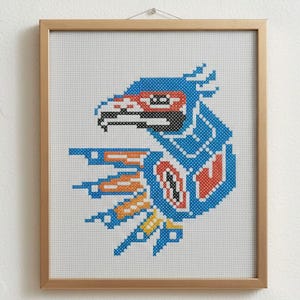 Puede incluir: Una obra de arte en punto de cruz enmarcada con un diseño de águila estilizada. El águila está representada en azul, naranja, rojo y negro sobre un fondo blanco. El marco es de color marrón claro.