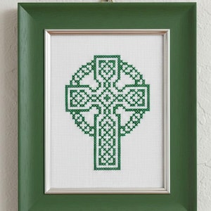 May include: A framed cross-stitch artwork featuring a green Celtic cross design. The cross is intricately stitched on a white background and framed in a green wooden frame with a silver inner border. The artwork is hung on a white wall.