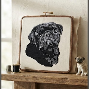 May include: A square wooden embroidery hoop containing a cross-stitch pattern of a black pug dog head. The design is stitched in shades of black and gray thread on a light beige fabric. A small metal thimble, a black thread spool, and a pug figurine.