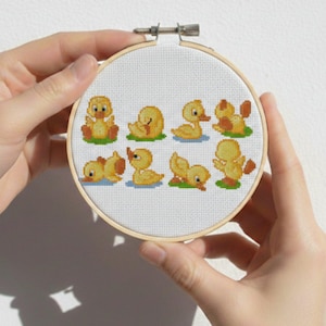 May include: A cross-stitch embroidery hoop featuring a pattern of eight yellow ducklings in various poses. The ducklings are stitched on white fabric with green and brown accents, set within a wooden hoop. The design is held by two hands.