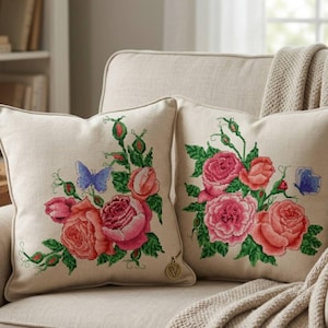 Può includere: Due cuscini quadrati con ricami floreali. I cuscini presentano un tessuto beige simile al lino con rose ricamate a punto croce nei toni del rosa e del corallo, foglie verdi e una farfalla blu. I cuscini sono su un divano beige.