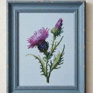 May include: Cross-stitch artwork of a purple thistle flower and bud with green leaves, set against a light blue background. The framed piece is in a distressed blue wooden frame, adding a rustic touch to the floral design.