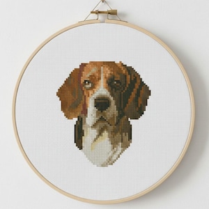 May include: A cross-stitch portrait of a beagle dog, with brown, white, and black pixelated details. The artwork is mounted in a round wooden embroidery hoop. The dog's face is centered, showcasing its distinctive markings. The hoop has a diameter of approximately 20 cm.
