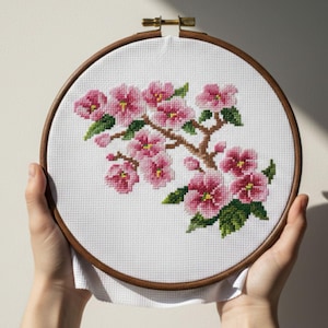 Könnte beinhalten: Kreuzstich-Stickerei eines blühenden Zweigs mit rosa Blüten und grünen Blättern. Die Stickerei befindet sich in einem Holzreifen, der von Händen gehalten wird. Das Design zeigt einen braunen Zweig und gelbe Zentren in den Blüten.