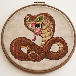 Op de afbeelding: Een kruissteekborduurwerk van een bruine cobra met groene ogen, een wijd opengesperde bek en een roze gespleten tong, gepresenteerd in een ronde houten borduurring. De slang heeft gedetailleerde schubpatronen in bruin- en tinten op lichte stof.