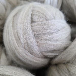 May include: A round ball of light beige, soft, fibrous yarn. The texture appears fuzzy and thick, with individual strands wound together in a dense, spherical shape. The color is a neutral, pale tan, consistent throughout the entire surface.