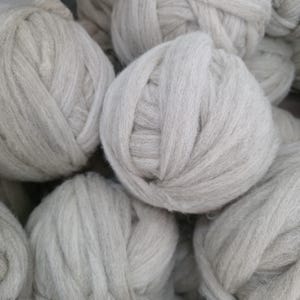 May include: A collection of multiple balls of thick, unspun wool roving in a light, neutral off-white or cream color. Each ball features a soft, fibrous texture with visible strands of natural wool fiber wound into a dense, rounded shape.