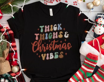 Puede incluir: Camiseta negra con el texto "Thick Thighs & Christmas Vibes" en letras de colores de estilo retro.