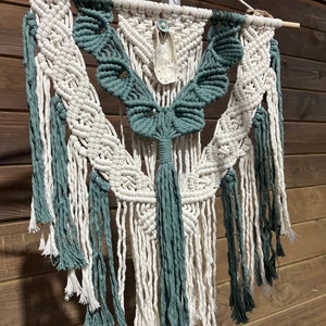 May include: A macrame wall hanging in cream and teal. The design features a central teal butterfly shape with cream-colored wings and hanging tassels. A decorative shell is attached at the top. The wall hanging is suspended from a wooden dowel.