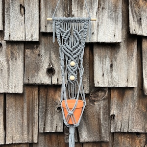 May include: A grey macrame plant hanger featuring intricate knotted patterns, two light-colored wooden beads, and a terracotta pot held within the cord structure. The hanger is suspended from a horizontal wooden dowel with a grey cord loop.