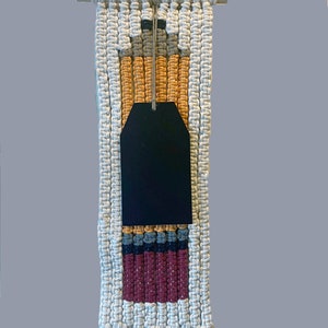 May include: A macrame wall hanging featuring a wooden dowel at the top with a hanging cord. The design incorporates off-white, mustard yellow, grey, and maroon knotted cord patterns. A large, blank black rectangular tag hangs from the center.