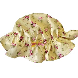 May include: A yellow floral sun hat with a wide brim. The hat features a pattern of pink and red roses with green leaves. A small bow with yellow and pink ribbons is attached to the front of the hat.