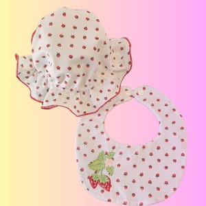 May include: A white baby bonnet and bib set, patterned with small red strawberries. The bonnet has a ruffled edge and red trim. The bib features a larger strawberry design, perfect for a baby.